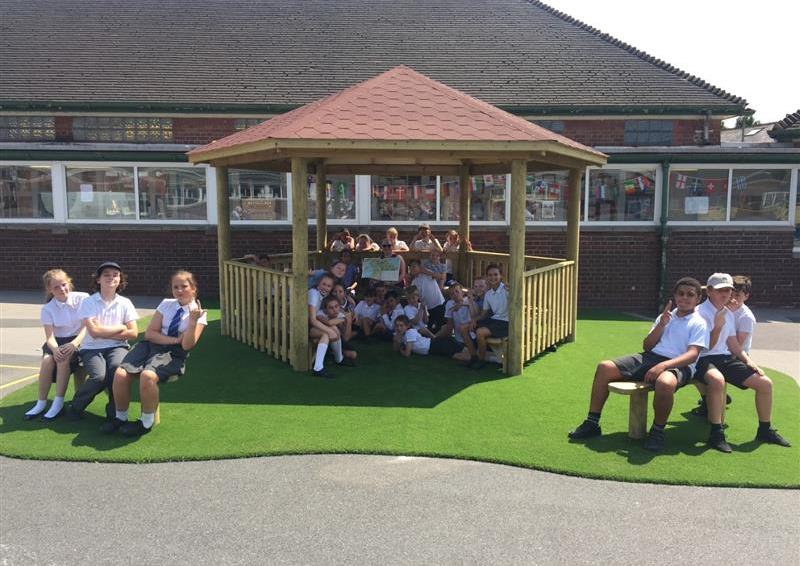 A wooden canopy that has a surface of artificial grass. The artificial grass has been installed on a concrete playground. A huge group of children are in the canopy, with a small group of children on each side of the canopy sitting on benches.