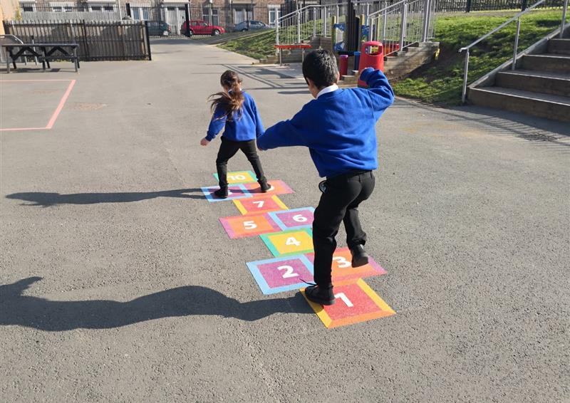 2 primary school children are playing on a thermoplastic playground marking of a hopscotch graphic. The thermoplastic graphic has been installed on a tarmac surface,