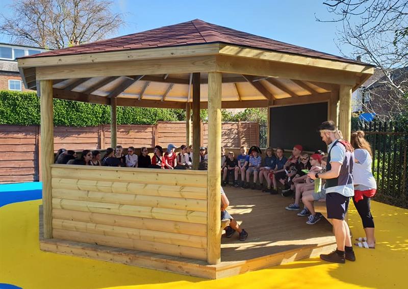 A wooden gazebo that has been installed on top of yellow wetpour. A huge group of children are sat all around the gazebo as two teachers stand at the entrance. The teachers are giving a lesson to the kids as they all look at the teachers.