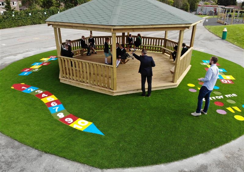 A wooden Gazebo that has been installed on an artificial grass surface and has Saferturf graphics around the Gazebo. The Gazebo currently has a group of children inside it and 2 teachers leading a lesson. Trees and natural grass fields can be seen in the background.