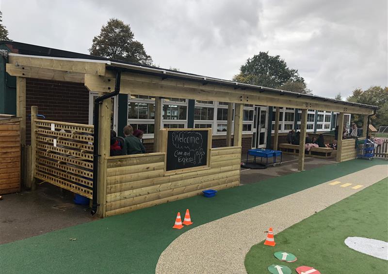 A wooden canopy has been installed on a wetpour surface. The canopy has been connected to a school, with a variety of outdoor play equipment stored underneath it. The canopy is huge, with a chalkboard installed on one of the walls.