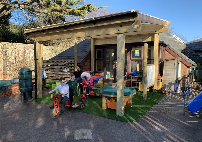 A timber canopy that has been attached to a school. A small area of artificial grass can be seen underneath the canopy, with a group of children playing underneath the canopy. A variety of different outdoor learning and play equipment.