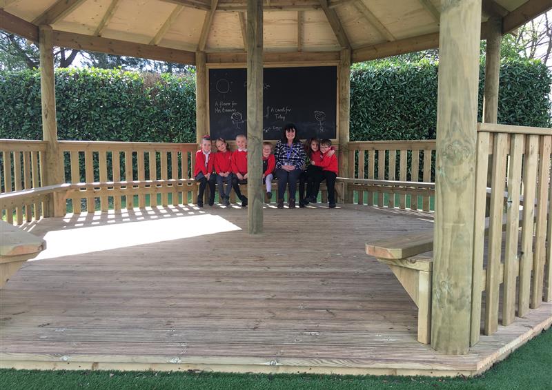 A group of children are sat inside a wooden gazebo, with a teacher sat in the middle. A wooden bench can be seen going around the entire gazebo. A clear view of the wooden flooring can be seen and a giant chalkboard can be seen behind the group of students and teacher.