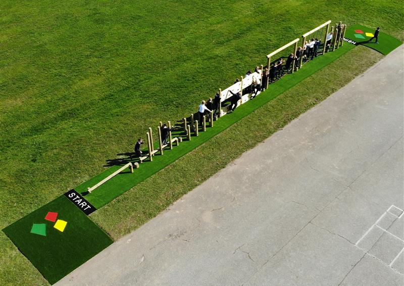 A birds-eye shot of a Trim Trail. The Trim Trail has been installed on artificial grass, which has also been installed on top of natural grass. The Trim Trail has a large group of children playing on the equipment, moving from the left to the right. There is also a variety of Saferturf graphics.