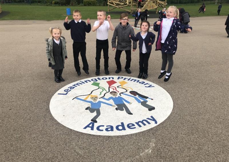 A bespoke playground marking that is a white circle, with markings of children holding kites in the middle. The text "Leamington Primary Academy" can be seen going around the edge of the circle. A group of children are stood above the graphic, smiling and waving at the camera.