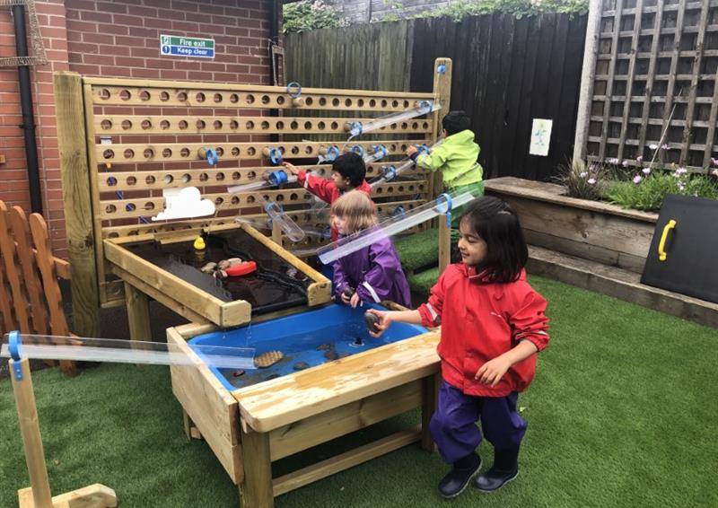 A group of children are playing and engaging with a variety of water play equipment. A water table, water dam and a water wall can be seen on the photo, all connecting to each other. The equipment is installed on an artificial grass surface.