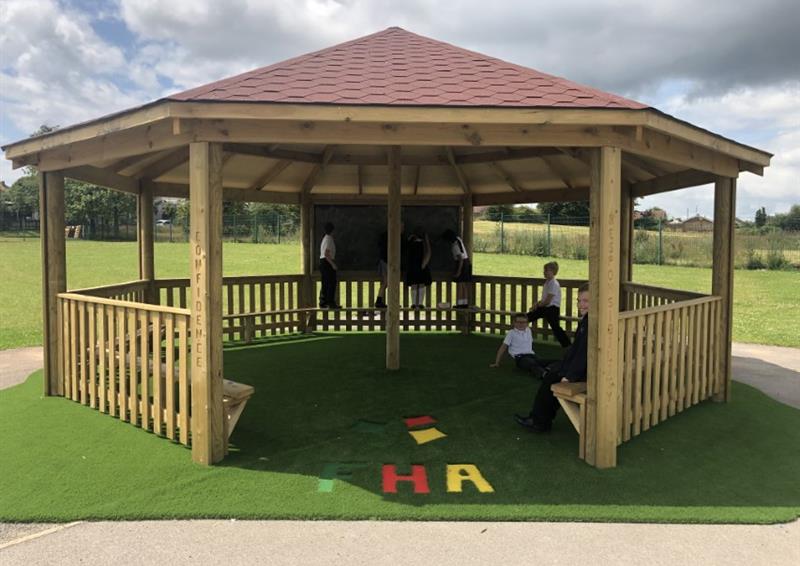 A wooden gazebo installed on an artificial grass surface. The Gazebo is an Octagon shaped, with a red roof.  On the inside, some children are playing with some outdoor play equipment. A natural grass turf surrounds the Gazebo, with trees in the far distance.