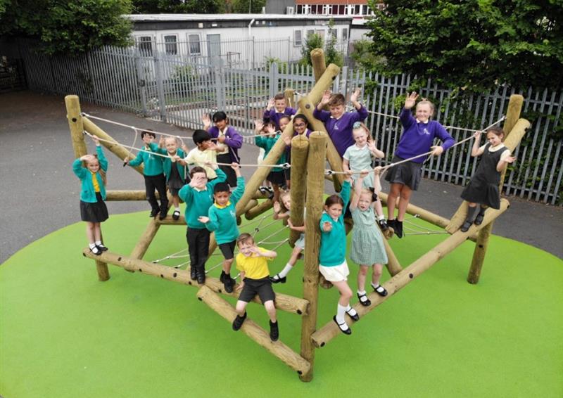 A climbing frame that has been installed on top of green wetpour on a playground. A group of children are playing on the climbing frame as they look at the camera, smiling.
