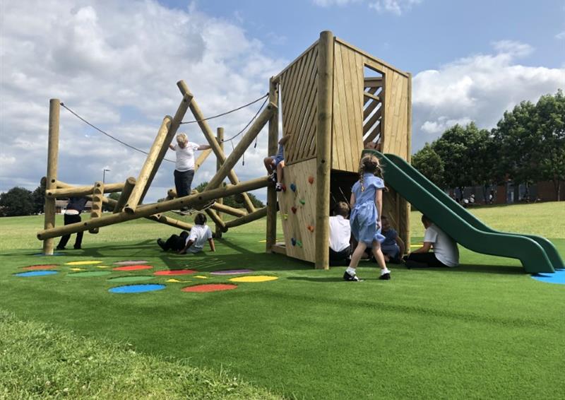 A climbing frame and a play tower are installed on artificial grass right next to each other. A group of kids are playing on both pieces of equipment. A few Saferturf graphics can be seen on the artificial grass.