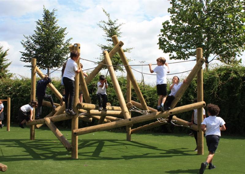 A climbing frame has been installed on an artificial grass surface. A group of children are climbing all over the equipment. Trees and bushes can be seen in the background.