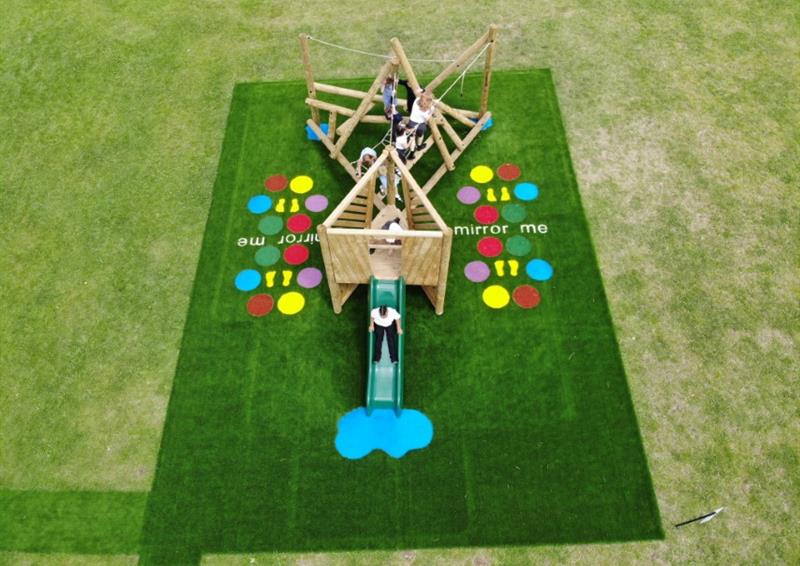 A birds-eye photo showing a climbing frame and a play tower being installed on the same patch of artificial grass. Both of these pieces of play equipment are installed right next to each other. A group of children are playing on the equipment.