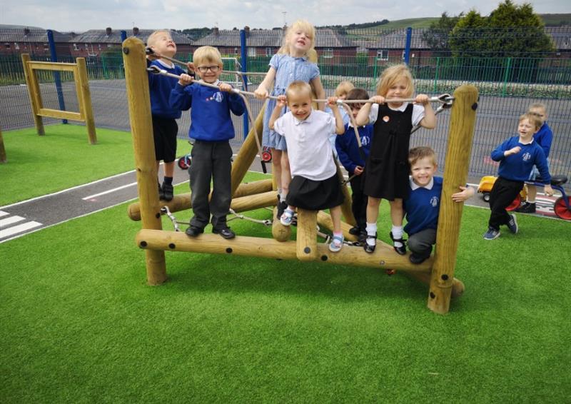 A Playframe has been installed on artificial grass, with a wetpour road going around the area. A group of children are playing on the climbing frame, as other children are running over to it.