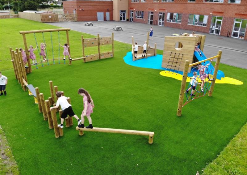 A class of children playing on different pieces of equipment on the trim trail, the trim trail is in a circle pattern and installed on top of a layer of artificial grass.