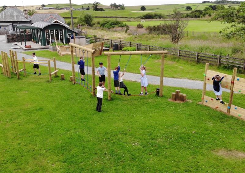 7 children playing on our trim trail, 1 young boy is standing to the side of the trim trail watching the other children playing. The Trim Trail is installed on natural grass, with a tarmac path behind the Trim Trail leading to the school.
