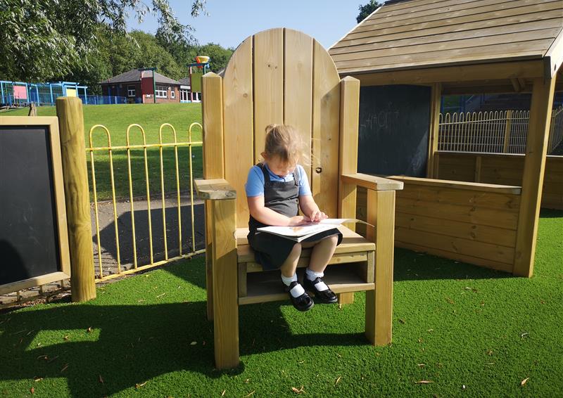 A little girl is reading a book whilst she sits in a big, wooden chair. The girl is a lot smaller than the chair, with her completely off the ground. The chair has been placed on artificial grass and next to a playhouse and a chalkboard.