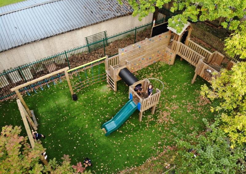 A play tower with a trim trail attached to it. The equipment has been installed on a natural grass surface. Some children can be seen playing on the equipment. A school building can be seen behind the equipment.