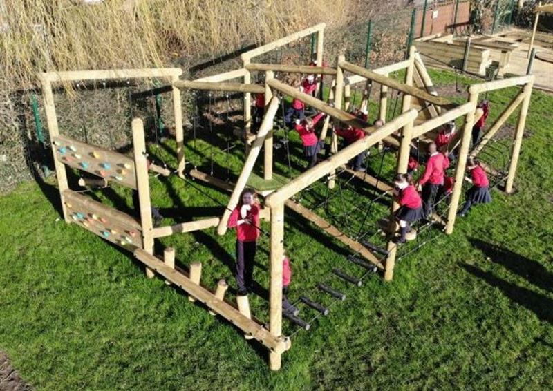 A Forest Circuit has been installed on natural grass, with a group of children playing on it. Trees can be seen in the background, with a group of planters being seen in the far distance.