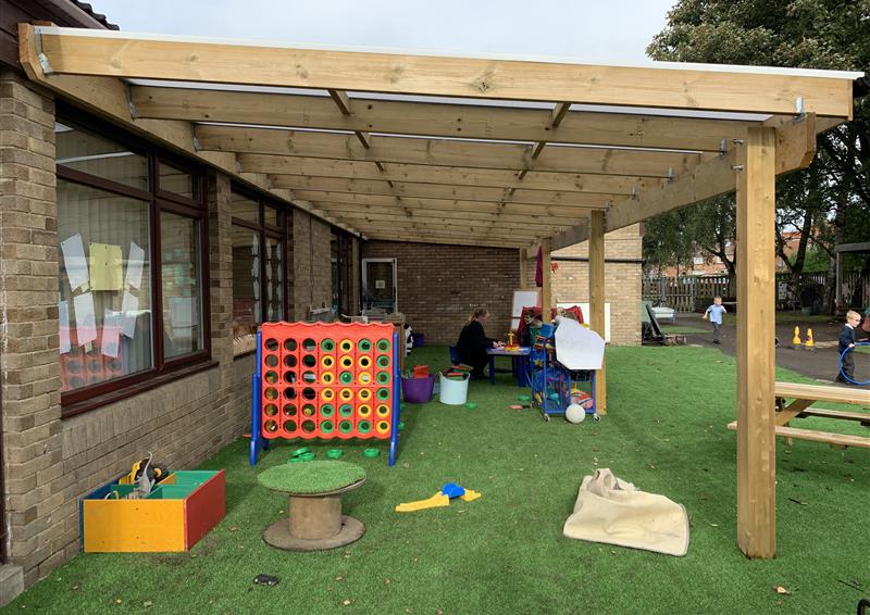 A timber canopy is attached to a school building. A wide variety of outdoor toys and learning equipment can be seen underneath the canopy. The surface underneath the canopy is artificial grass.