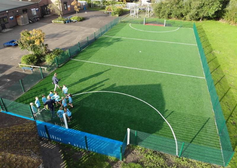 A green artificial grass MUGA surface, with a green fence going around the entire pitch. A group of kids can be seen on one side of the pitch, grouping around the goal. The goals have white posts and have either a blue or red wetpour surface.