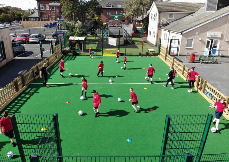 A MUGA Pitch with a dark green colour and a mixture of wooden and metal fencing. A group of children are playing with footballs on the pitch as they dribble the ball around cones. The goals are built into the metal fencing.