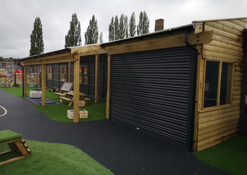 A timber canopy is built on top of a surface of wetpour and artificial grass. The canopy has a metal shutter to protect outdoor equipment. It also has windows and walls, allowing for classes to use the space for learning.