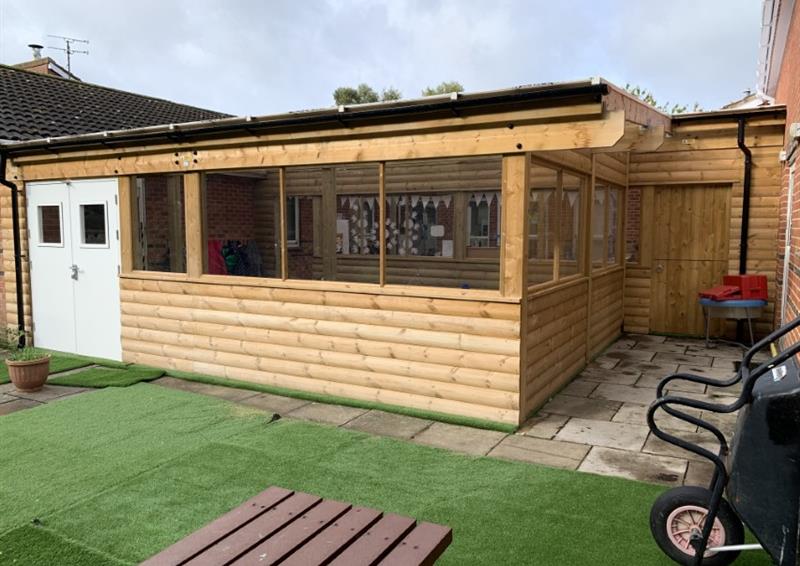 A wooden canopy installed on an artificial grass surface. It has double white doors on the entrance, with a row of windows going around the whole structure. A variety of outdoor equipment can be seen scattered around the play area that lies in front of the building.