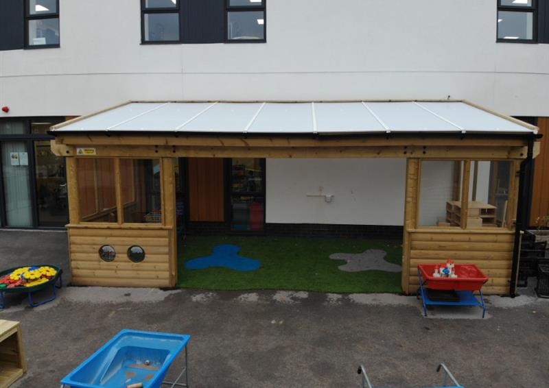 A wooden canopy is installed on top of a concrete playground, with an artificial grass surfacing installed inside the structure. The canopy is connected to the school and has a white roof, with windows and walls, and has a big entrance at the front.