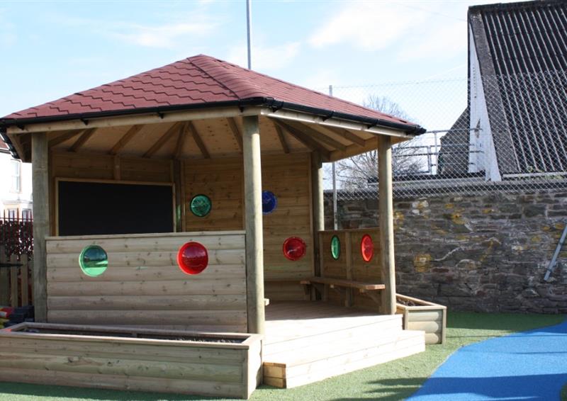 Wooden gazebo that has been installed on green wetpour. The walls of the gazebo have circles cut out of them, with them being coloured red or green. The roof of the structure is red, with planters being placed on the left and right of the entrance to the Gazebo.