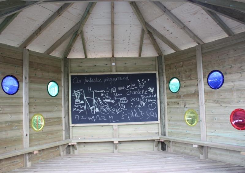 The inside of a wooden gazebo. The back wall has a chalkboard attached to it, with the rest of the walls have 2 circles cut out on each panel. Over each circle is a coloured piece of plastic, such as red, blue, yellow, green.