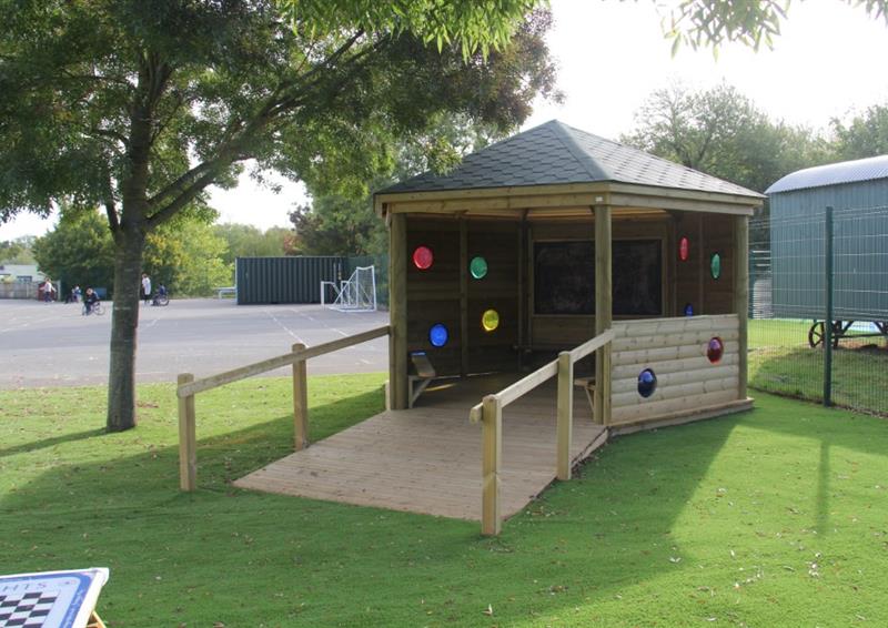 A wooden gazebo with a wooden ramp leading to the entrance of the structure. A variety of circular windows can be seen installed on the side of the Gazebo, with coloured plastic covering the holes. The colours are red, green, blue and yellow.