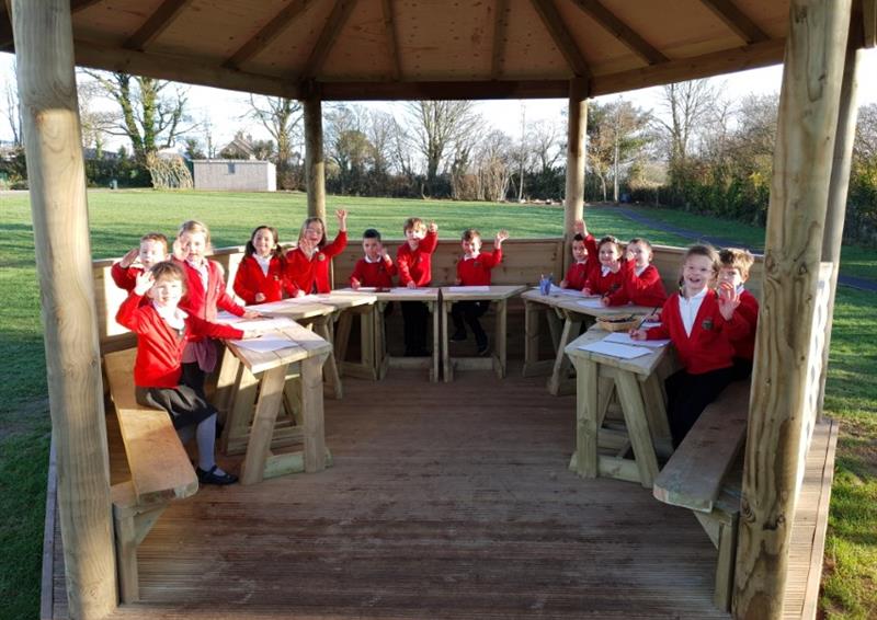 A group of children are sat on benches that are attached to the inside of a wooden gazebo. They all have small tables in front of them with work placed on them. The children are all looking at the camera and smiling. In the background, you can see trees and grass, creating a natural environment.