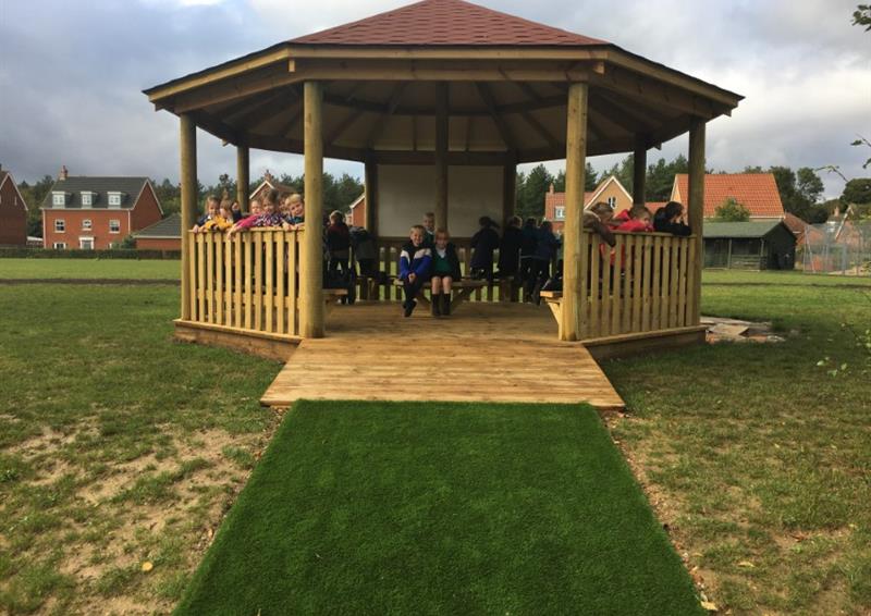 A wooden gazebo installed on natural grass, with a wooden ramp that connects the entrance of the structure to an artificial grass path. In the inside of the wooden gazebo, a group of children can be seen sat around and looking in different directions, taking in the nature that surrounds them.