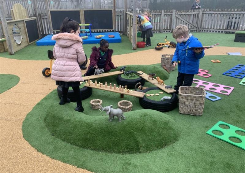 Artificial grass surface that has been installed in an EYFS setting. 2 artificial grass mounds can be seen in the photo with a group of children engaging with play equipment that is laid out on the surface.