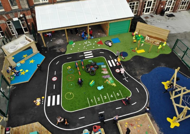 A variety of thermoplastic playground markings can be seen all around a playground. The most prominent one is a road marking, that goes in a circle on black tarmac. Artificial grass can be seen on the playground too.