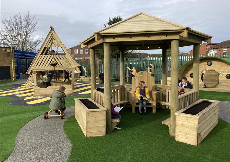 A tall, wooden Gazebo with a wooden chair in the centre. A child is sat on the chair, reading a book. A planter can be seen on both the right and left of the entrance to the Gazebo. A child is on a tricycle, riding past the Gazebo.