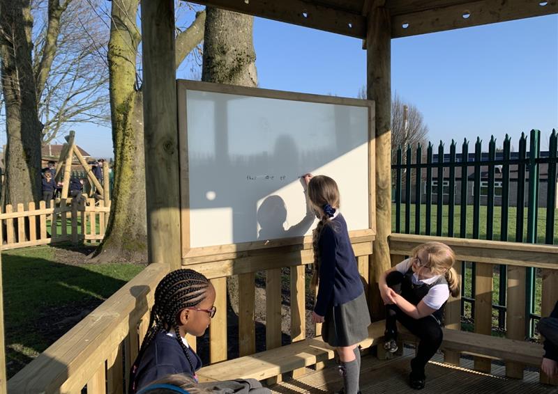 Three children are sat inside a wooden gazebo and are looking at a whiteboard that is placed at the back. One child has a marker and is writing on the whiteboard. You can see trees and grass in the background, highlighting the natural surrounding.