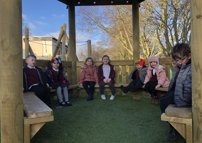 A group of children are sat on internal benches that are within a wooden gazebo. The children are all looking at the camera and smiling. Trees can be seen in the background, surrounding the kids in nature.