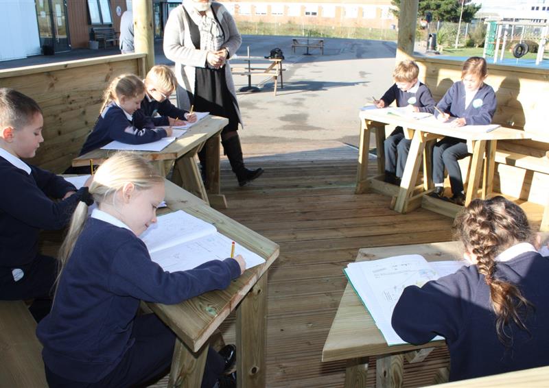A group of children are working at desks that have been set up within a wooden gazebo. The children are writing in their books as a teacher watches over them. The playground can be seen in the background, being a tarmac playground.