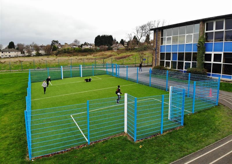 An artificial grass MUGA with a light-blue fence going around it. The court has built in football goals, with white posts. There are white and yellow line makings on the artificial grass.