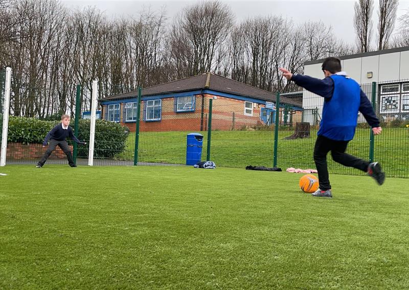 A Multi Use Games Area with an artificial grass surface. A child is taking a penalty kick whilst another child stands in goal. A green fence goes around the court, with the football goal's posts being coloured white.