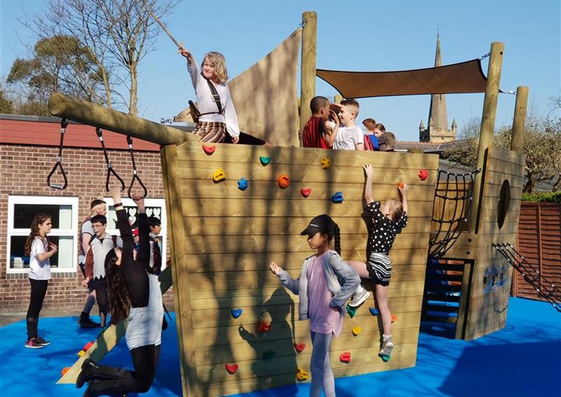 12 children playing on their new north sea galleon ship, 1 girl is swinging on the monkey bars, 1 girl is climbing up the climbing wall on the side of the ship and the rest of the children are playing onto or around the ship.