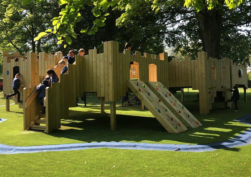 A class of children playing on the treehouse, 5 children are climbing up the stairs and 1 young girl is climbing on the climbing wall which is on the side of the treehouse.