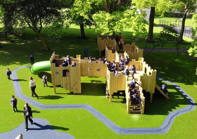 A class of children playing on their new bespoke treehouse, teachers are accompanying the children as they play. Artificial grass surrounds the bespoke treehouse.