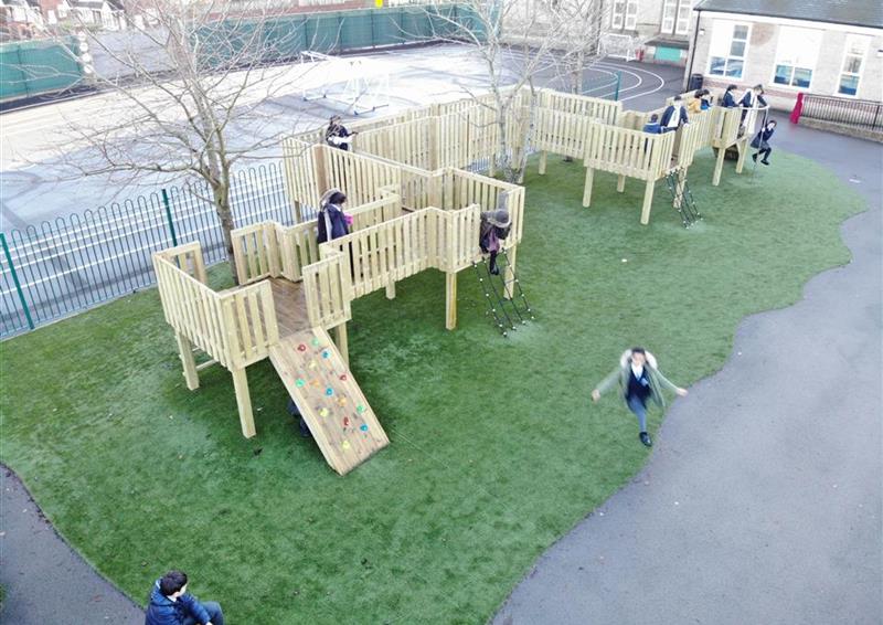 12 children playing on their new bespoke treehouse, 1 young boy is sat on the artificial grass next to the treehouse, 1 young girl is sliding down the pole on the side of the treehouse.