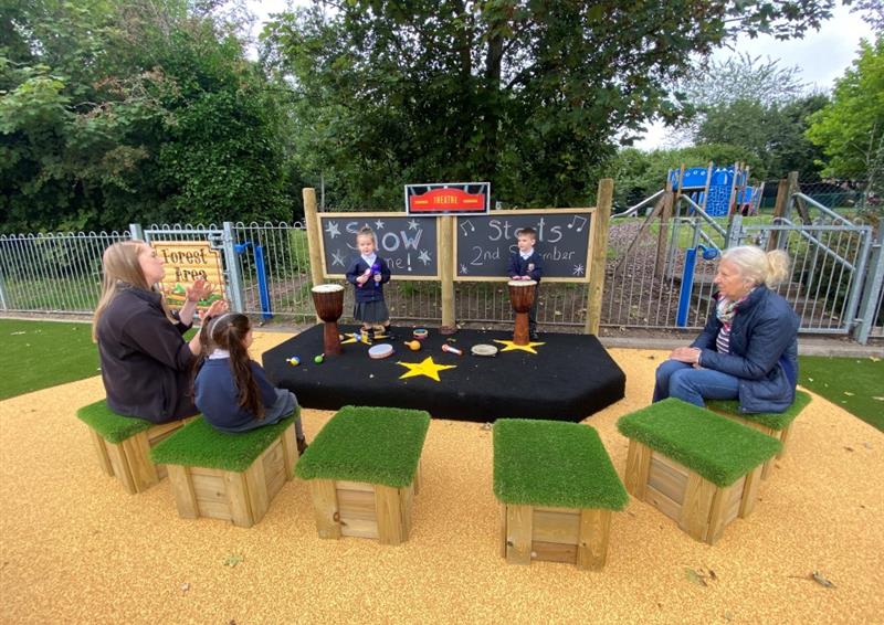 two adults and one young girl are sat on grass top seats in front of the performance stage. 2 children are playing drums on the performance stage.
