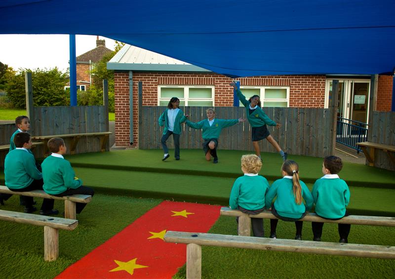 3 young girls dancing on the performance stage at their school, 6 other children are sat on the benches that are facing the stage with a red Saferturf carpet running through the middle.