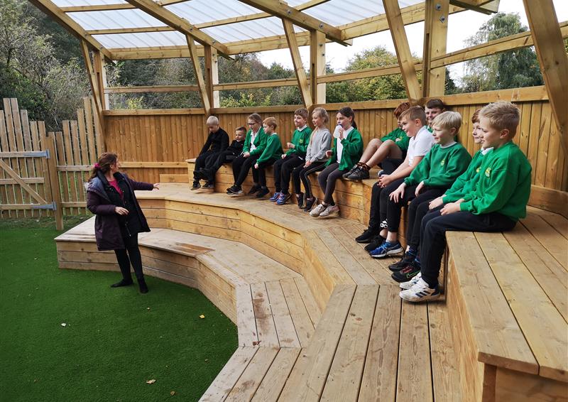 13 children sat in a row in our amphitheatre whilst the teacher is stood at the front and teaching a lesson.
