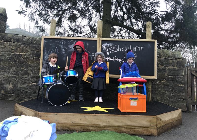4 children playing on their new performance stage, one young boy is playing on the drums and one young boy is playing the piano.