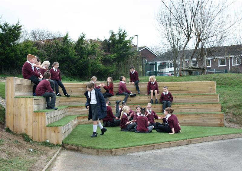 A class of children playing on their amphitheatre performance stage, artificial grass covers the top of the seats and the floor.