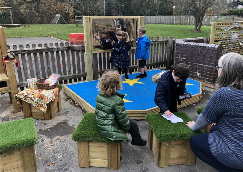 2 young children playing on their new performance stage drawing on the mirror in the background. 2 children and one adult are sat on the grass top seats in front of the stage.